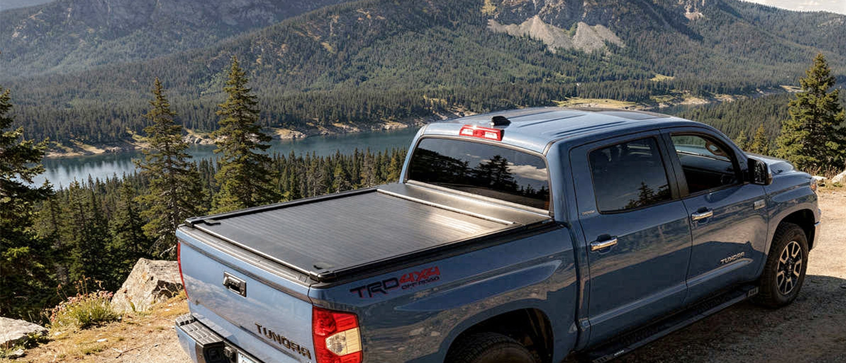 Blue truck with a tonneau cover parked on a scenic mountain road with a lake and trees in the background.