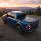 A blue Ford pickup truck with a black tonneau cover on the truck bed, parked on a dirt trail with mountains and trees in the background at sunset.