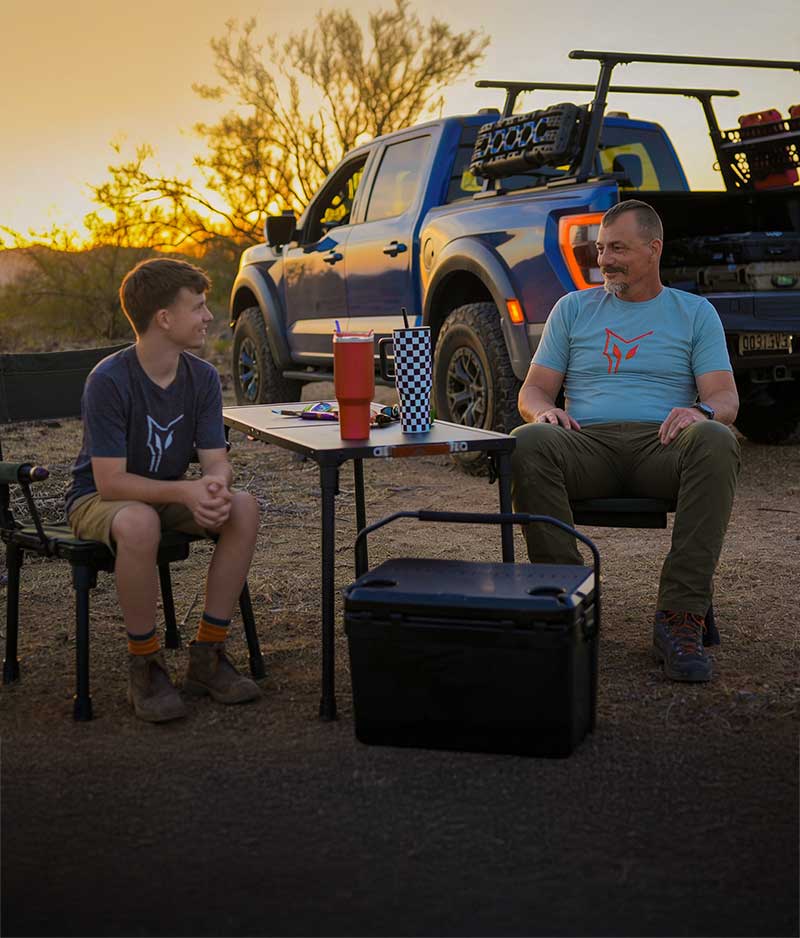 Two people sitting at a table outdoors with a blue truck in the background during sunset.