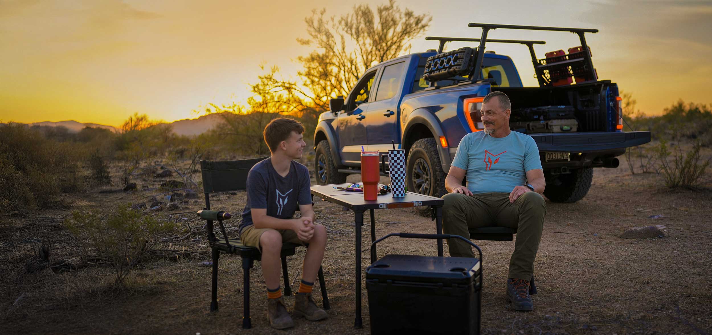Two people sitting at a table with a truck in the background during sunset.