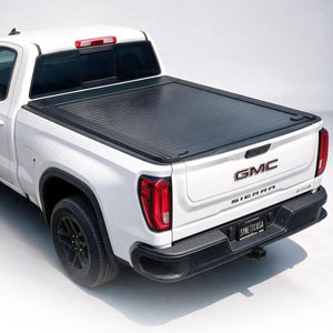 A black roll-up truck bed cover installed on the bed of a white GMC Sierra pickup truck, shown from a rear three-quarter angle on a clean white studio background.
