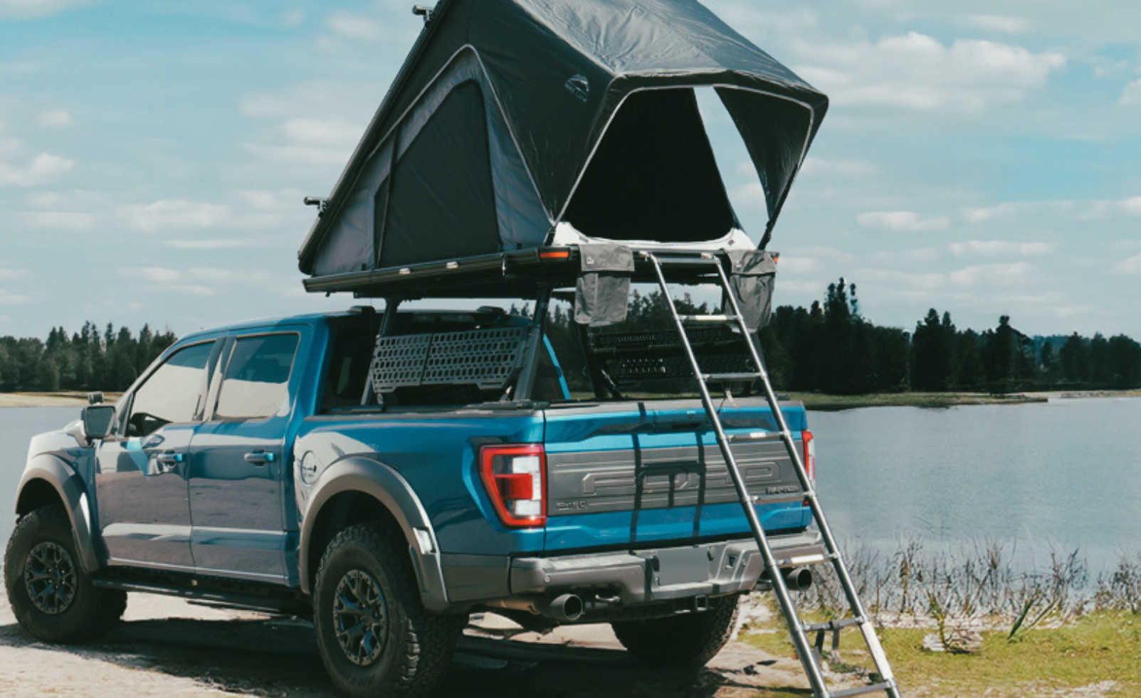 Blue pickup truck with a roof tent and ladder by a lake