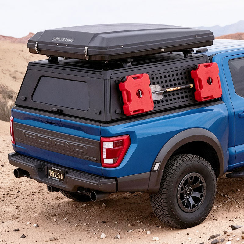An orange Ford pickup truck parked in a desert setting, shown from the rear three-quarter angle with a black roof cargo box and a black hard cap over the bed. Red side-mounted storage containers are attached to the bed, and the background shows a sandy desert landscape under a pale sky.