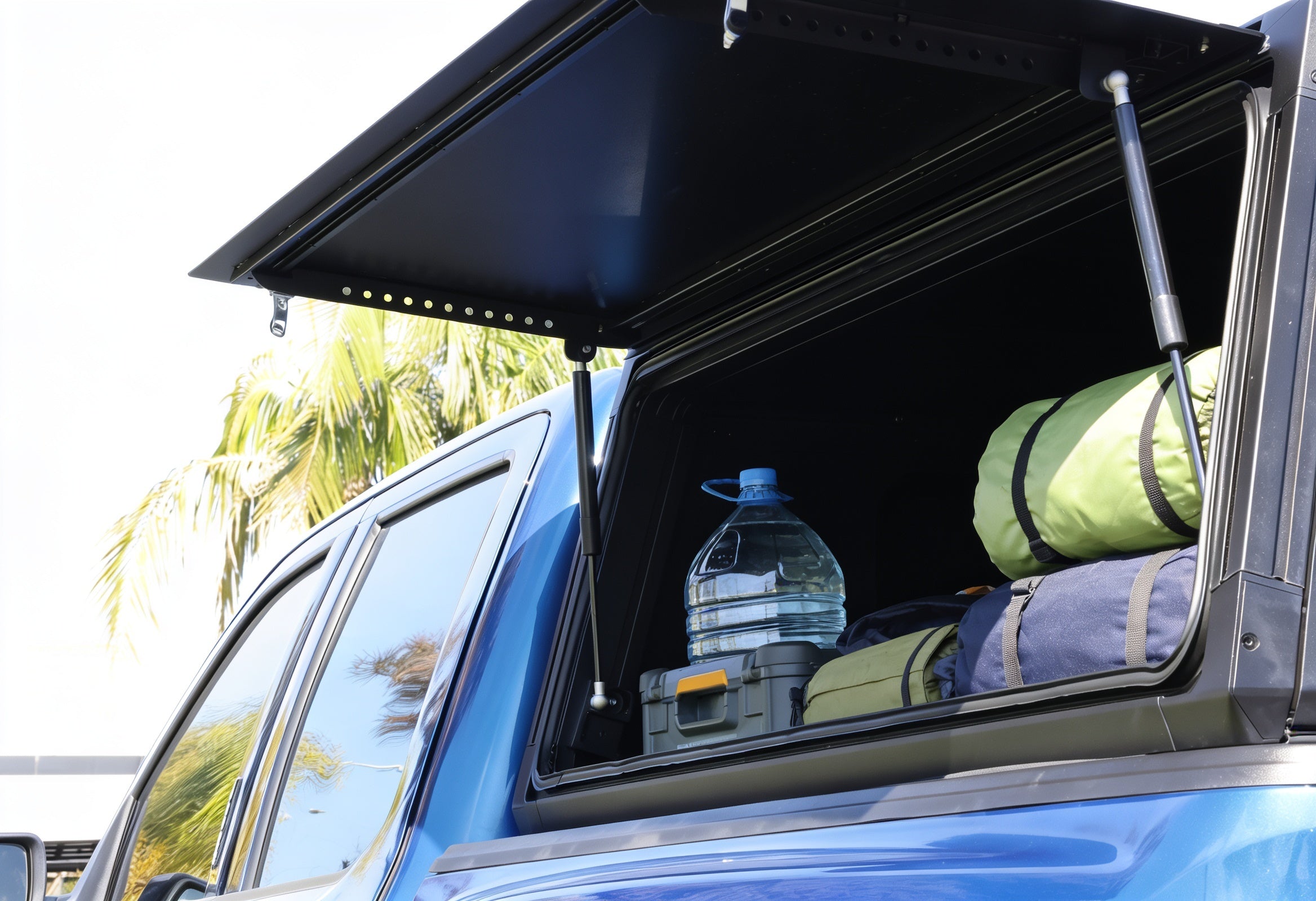 A black hardtop canopy mounted on the side of a blue vehicle, shown with its window open and loaded with camping gear including a green sleeping bag, a water jug, and a gray storage box. Palm trees are visible in the background.
