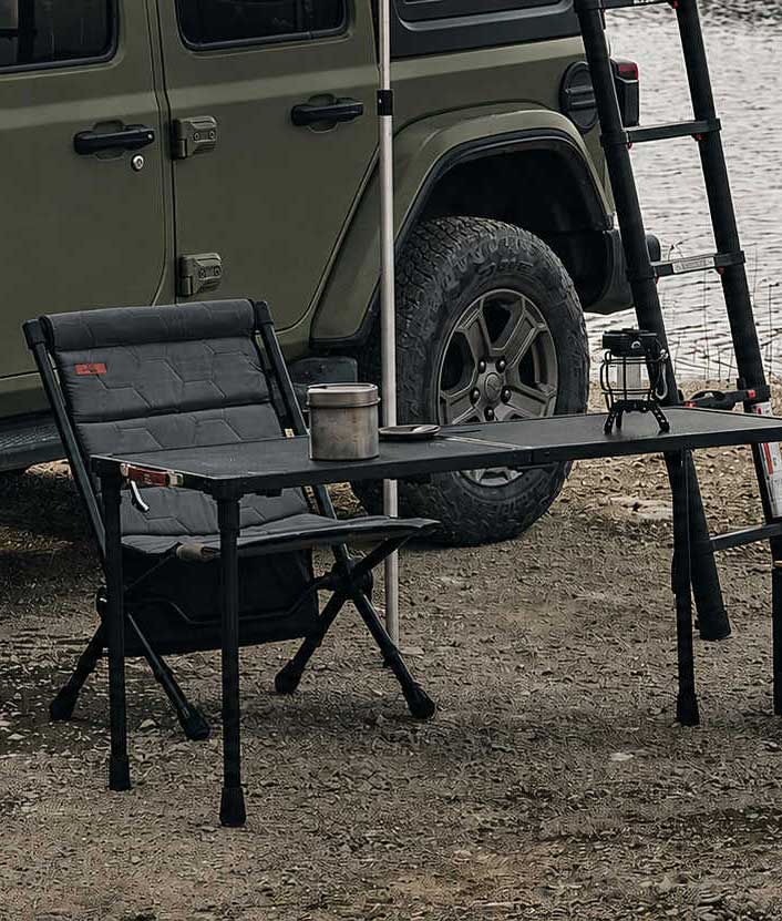 Outdoor setting with a green Jeep, folding table, and chair near a body of water.