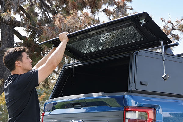 A man in a black t-shirt is shown lifting the lid of a black hardtop truck canopy mounted on the bed of a blue pickup truck, set against a background of trees and sky.