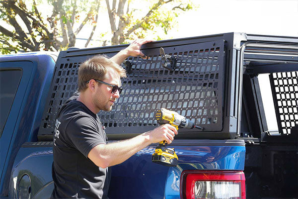 A man wearing sunglasses and a black t-shirt uses a yellow cordless drill to install a black metal mesh truck bed rack on the tailgate of a blue pickup truck, with trees and a bright sky in the background.